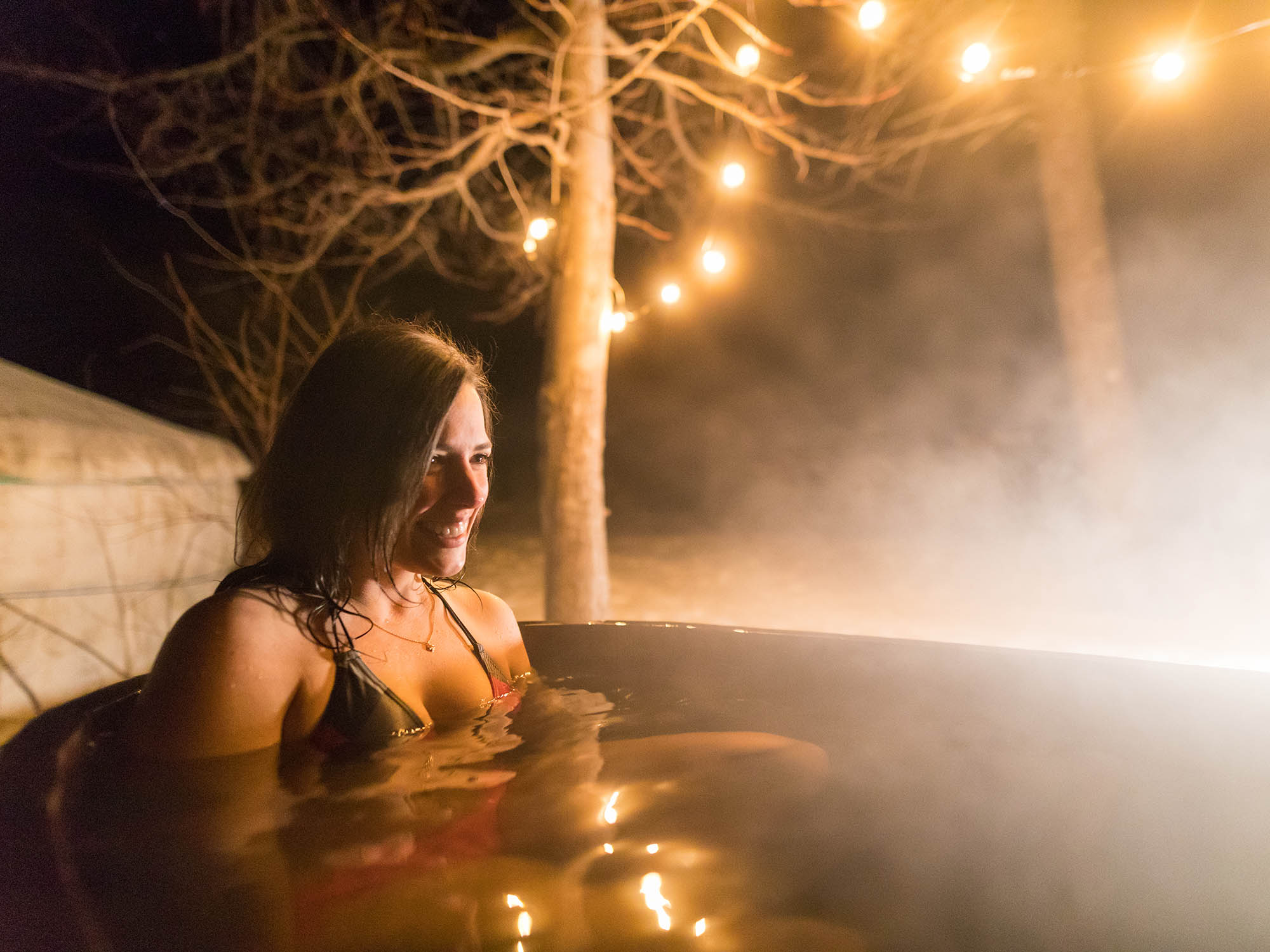 A person is smiling while sitting in a wood-fired hot tub at night at Cabot Shore Wilderness Resort on the Cabot Trail in Nova Scotia.
