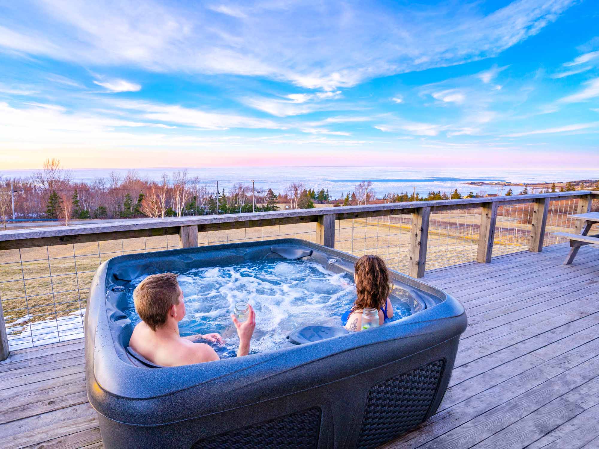 Two people taking in the view over the icy Northumberland Strait on a sunny winter day from the hot tub on the deck of one of the cottages at Steinhart Distillery in Arisaig, Nova Scotia.