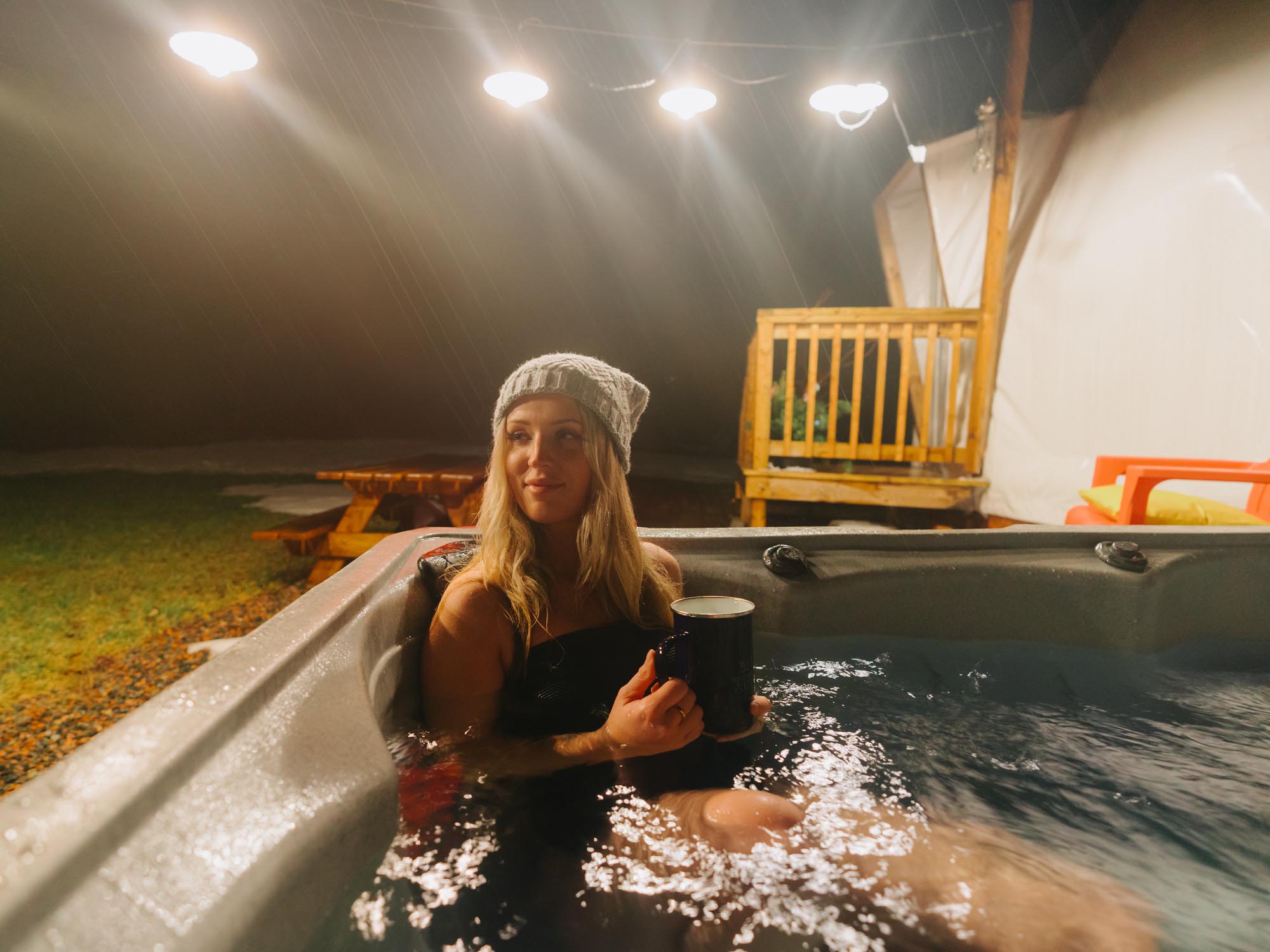 A person is enjoying the hot tub with a warm drink during their stay at Wallace River Ranch & Domes on a wintery night in Nova Scotia.