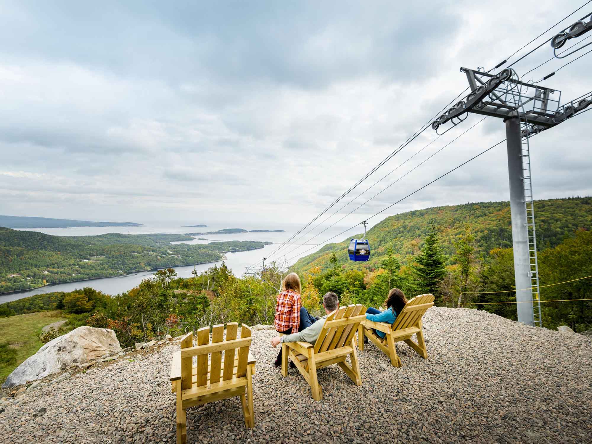 Family sitting in chairs at the top of Cape Smokey watching the gondola travelling to the top of the mountain with the early autumn colours around them and the amazing view over Ingonish in the distance in Cape Breton, Nova Scotia