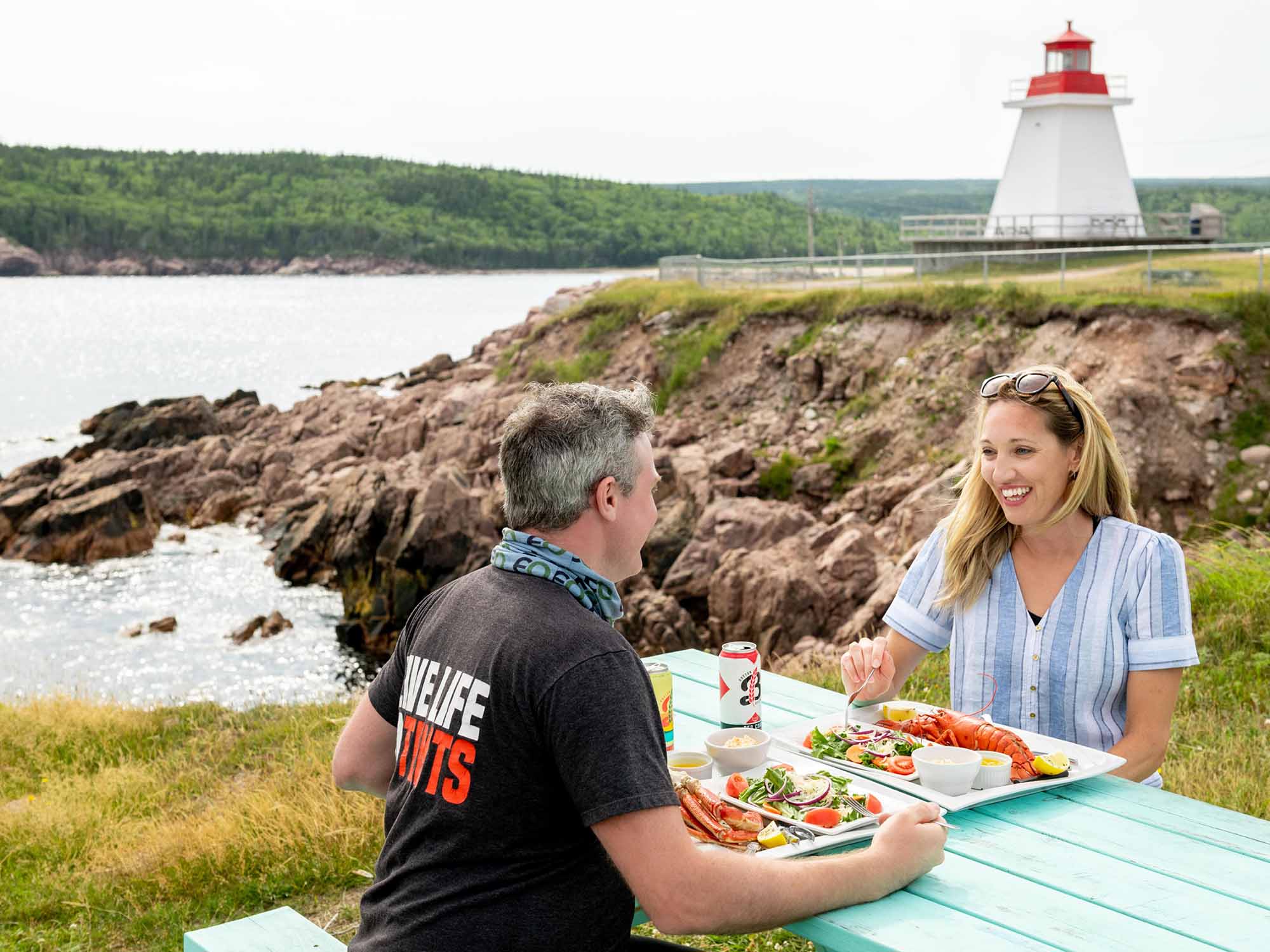 Two people eating lobster meals from the Chowder House in Neil's Harbour from a seaside picnic table with the Neils Harbour lighthouse in the background on the Cabot Trail in Cape Breton, Nova Scotia