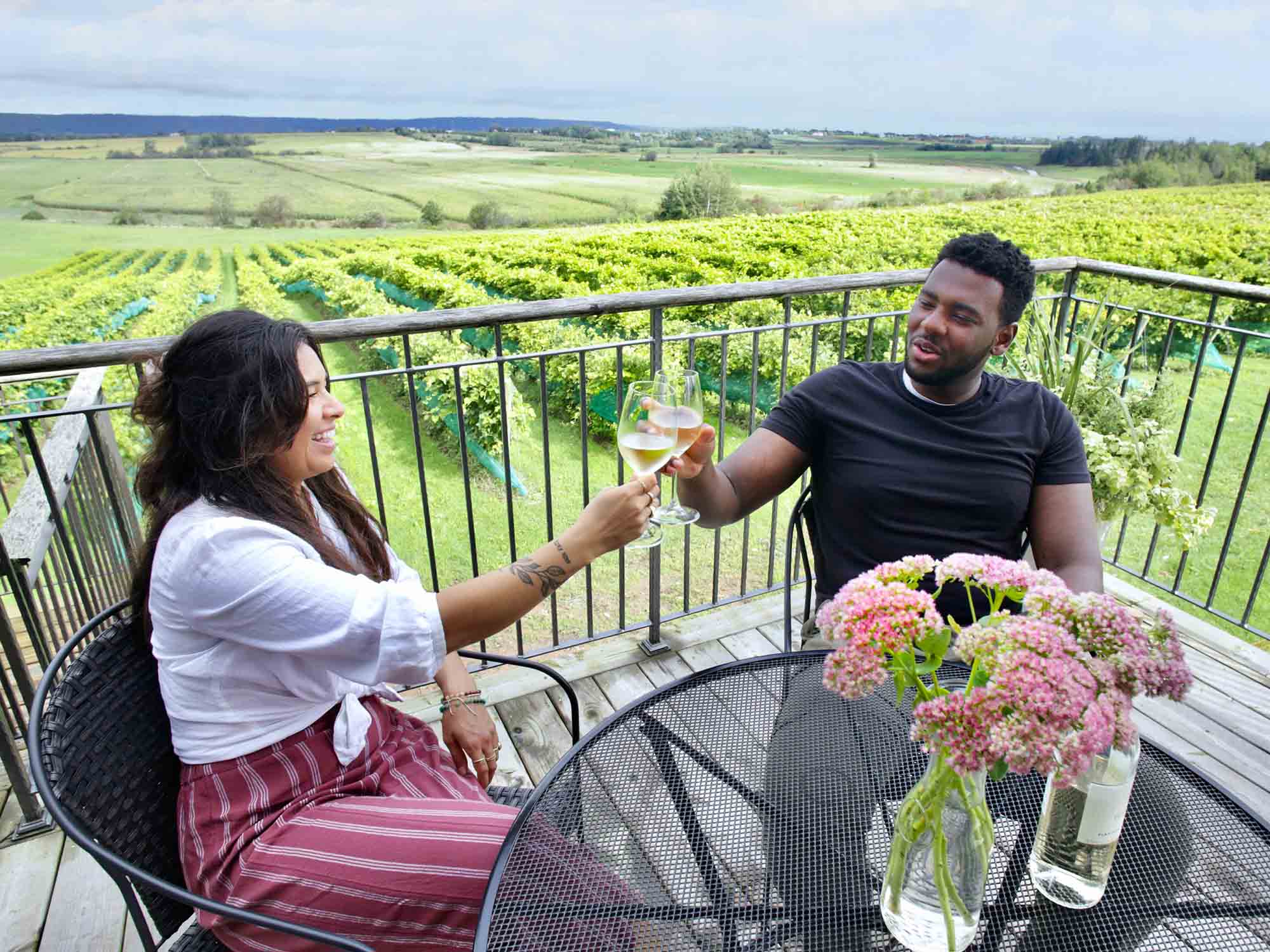 Two people doing a cheers with their glasses of Tidal Bay wine on the patio at Planters Ridge Winery with the lush vineyard behind them in Port Williams Nova Scotia