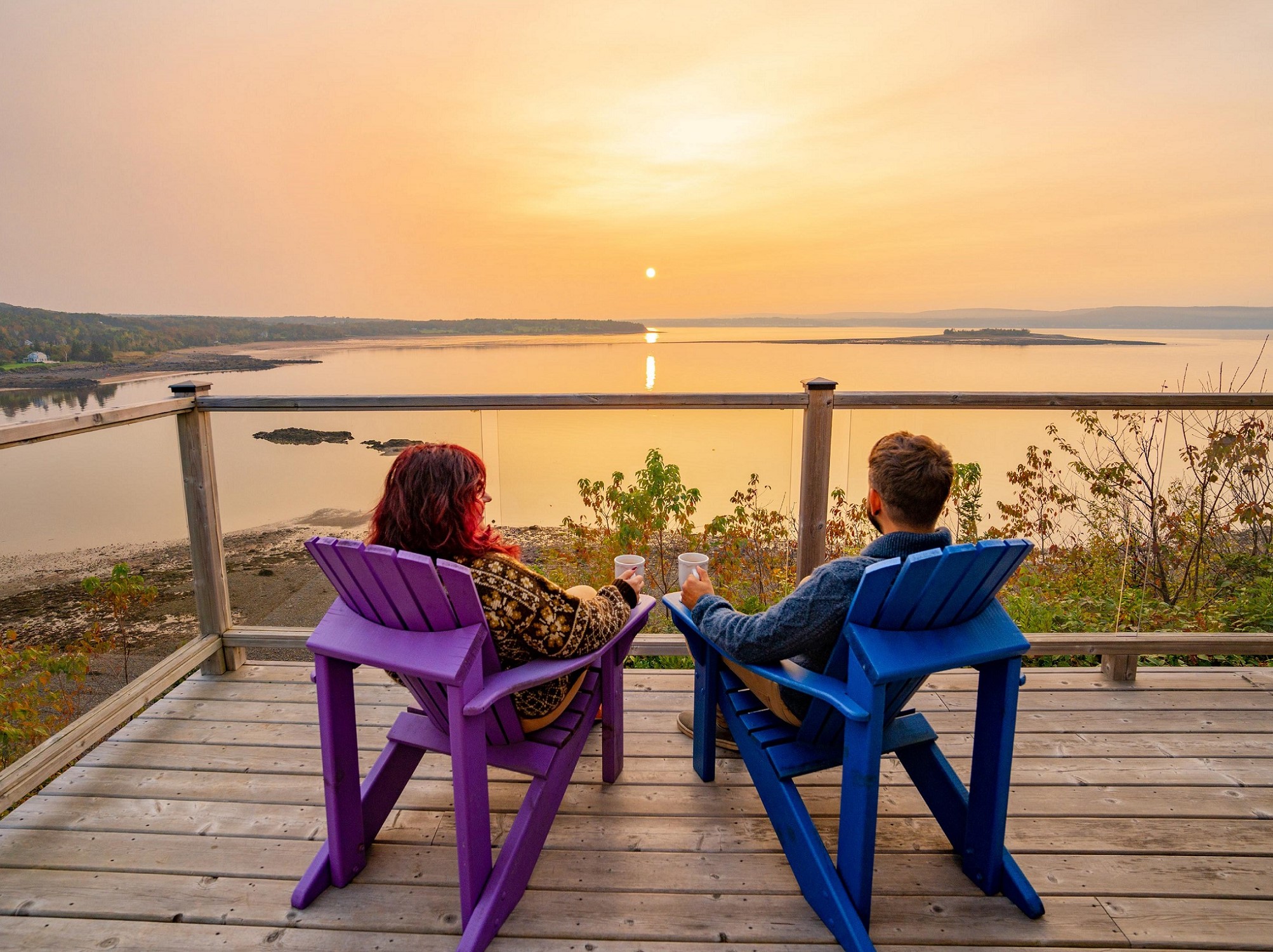 A couple sits in a purple and blue Adirondack chair with white mugs looking out over a golden sunset over the water from a deck.