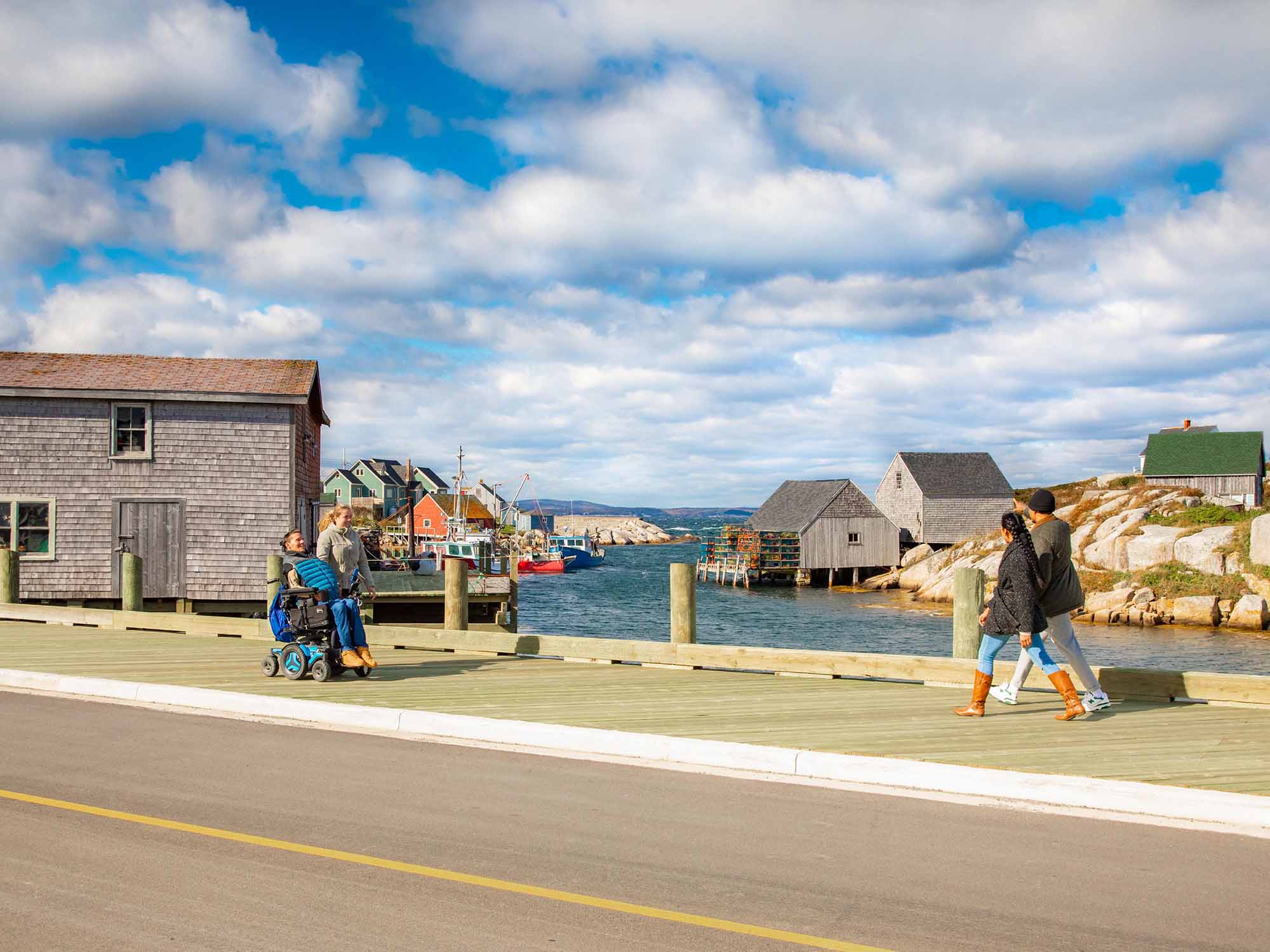 Four people, one in a motorized wheelchair, are walking on the new wheelchair accessible boardwalk sidewalk through Peggy's Cove village on a sunny fall day in Nova Scotia