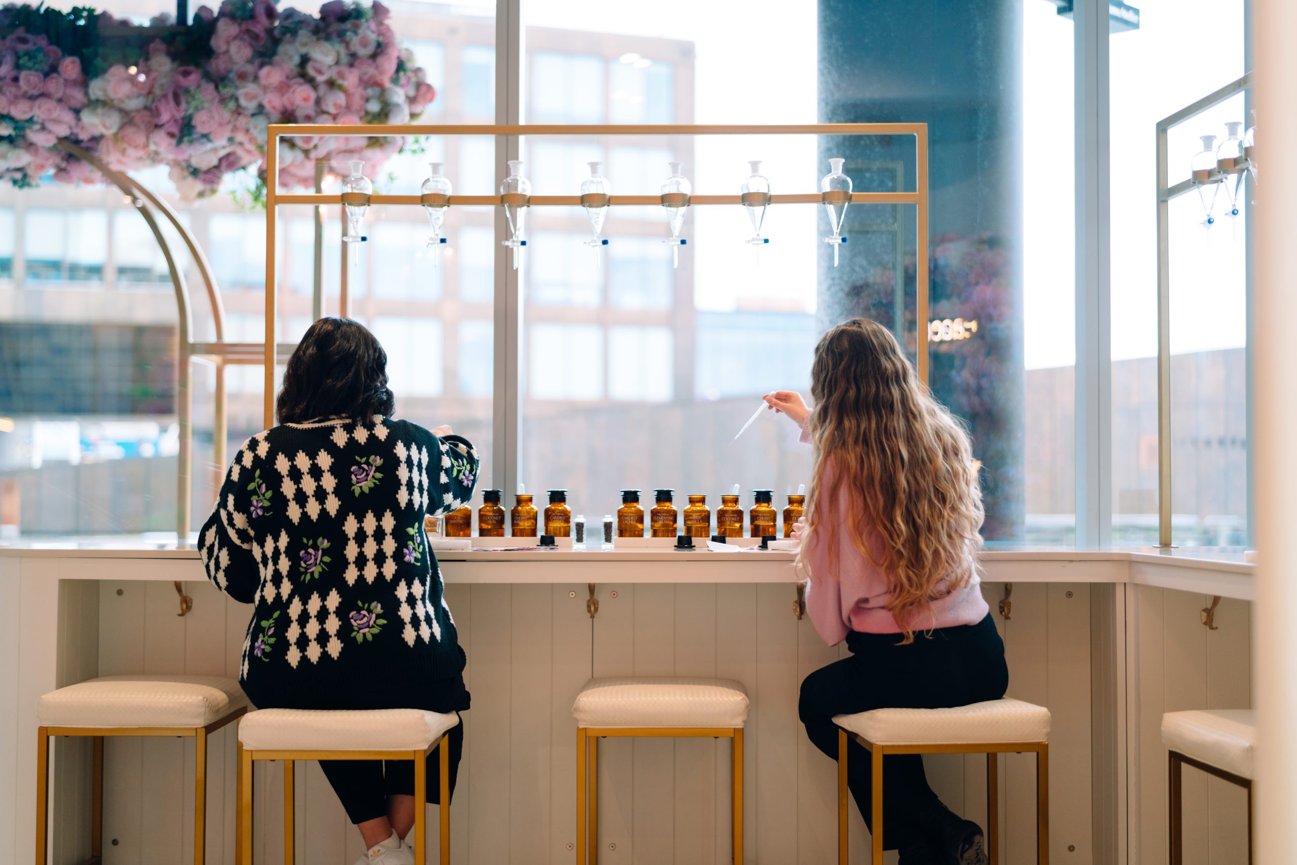 A woman in a black and white sweater and a woman in a pink sweater sitting on stools, in front of a window, making a custom perfume.