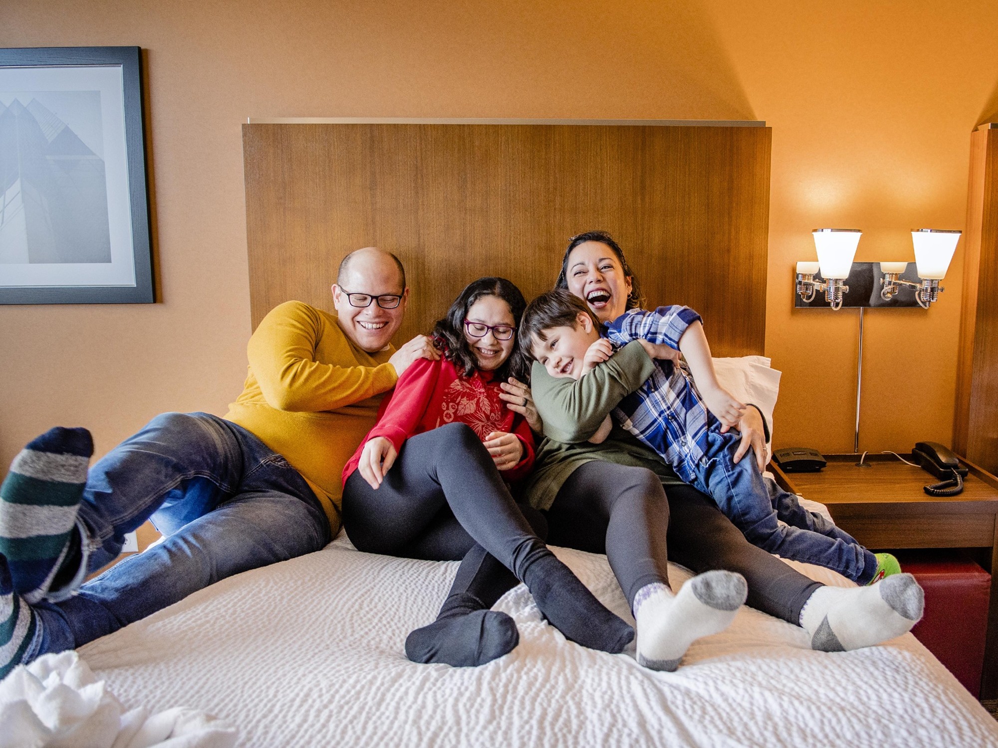 A family laughs together while sitting on a hotel bed.