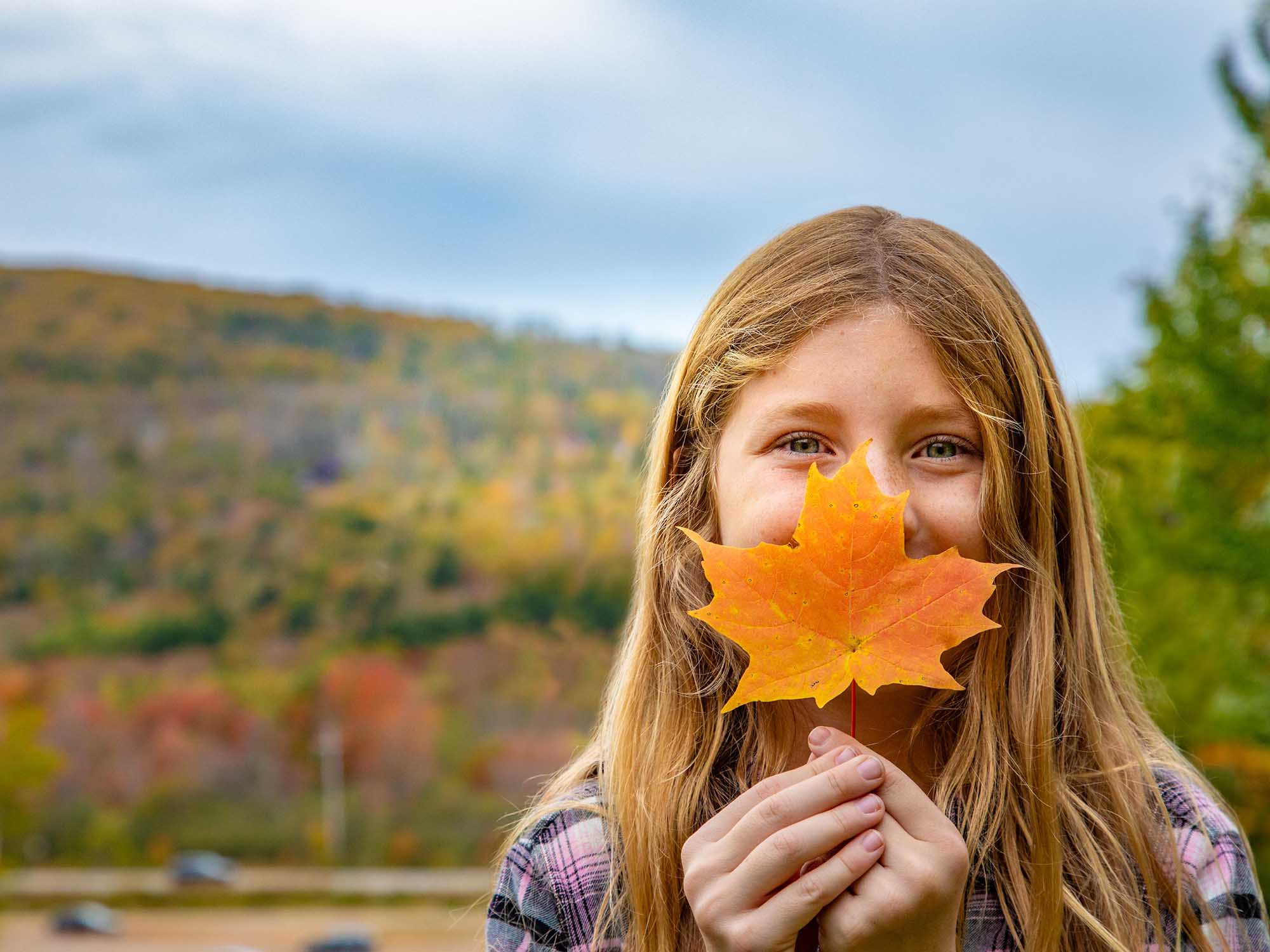 A young person holding an orange maple leaf in front of their face with the fall colours in the Wentworth Valley behind them on an autumn day in Nova Scotia