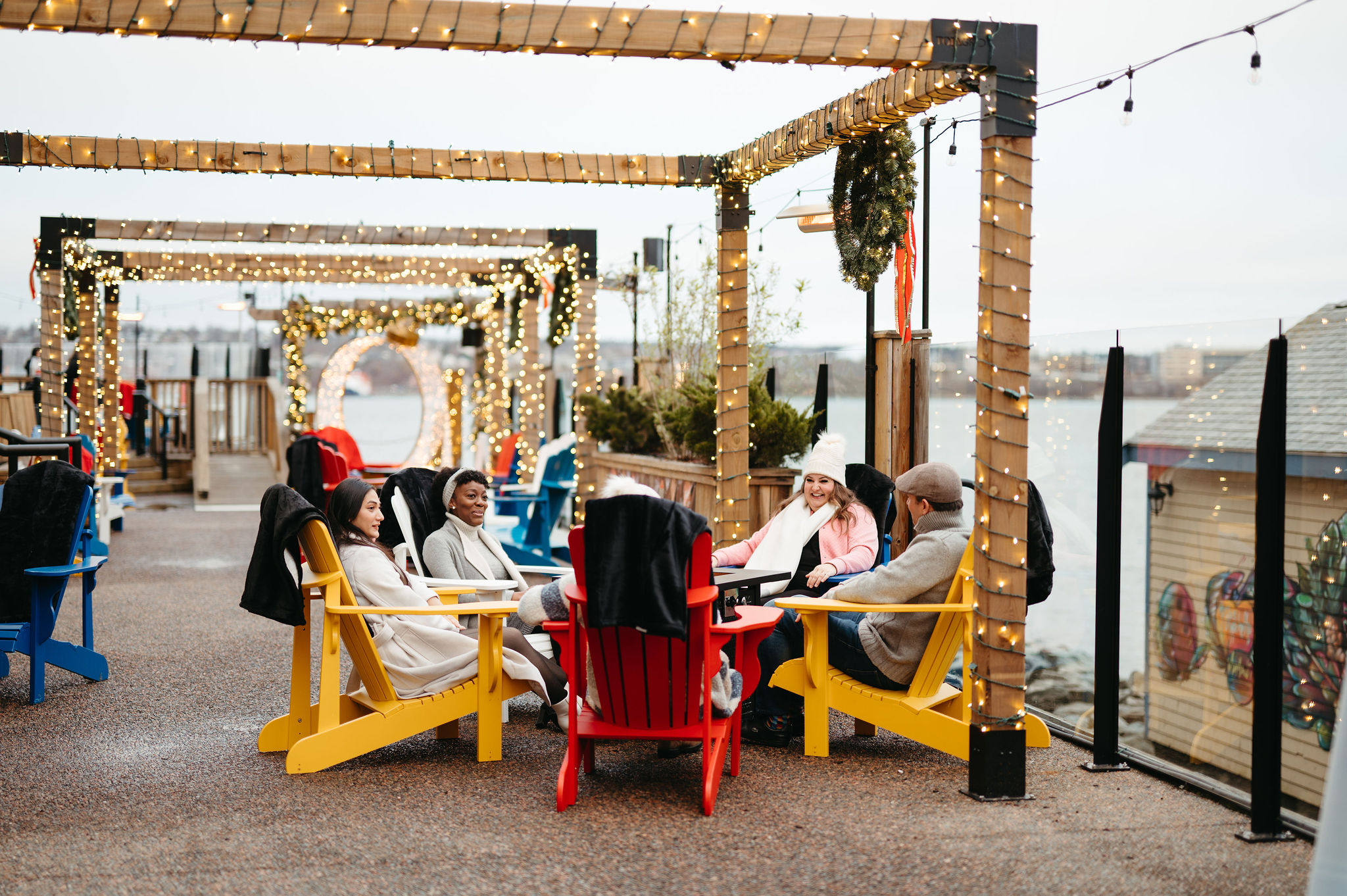 Friends sit around a fire in Adirondack chairs with Christmas lights overhead.