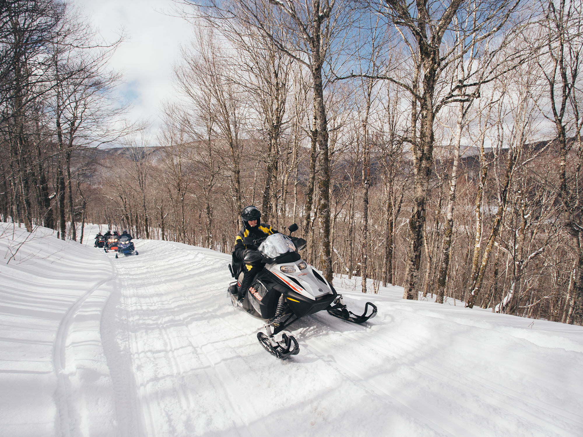 A group of individuals on snowmobiles on a trail surrounded by trees