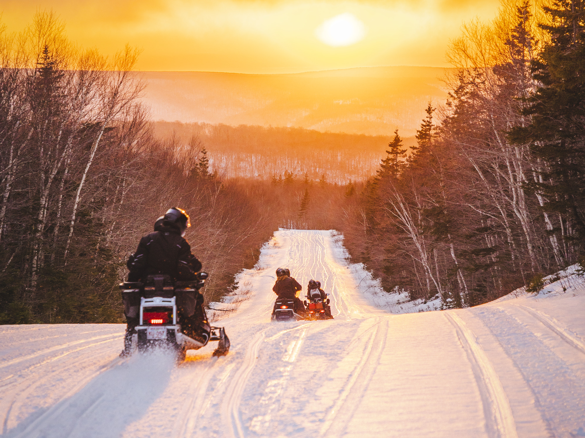 Snowmobilers on a snowy trail heading towards the winter sunset in the Cape Breton Highlands with TNT Outdoor Adventures.