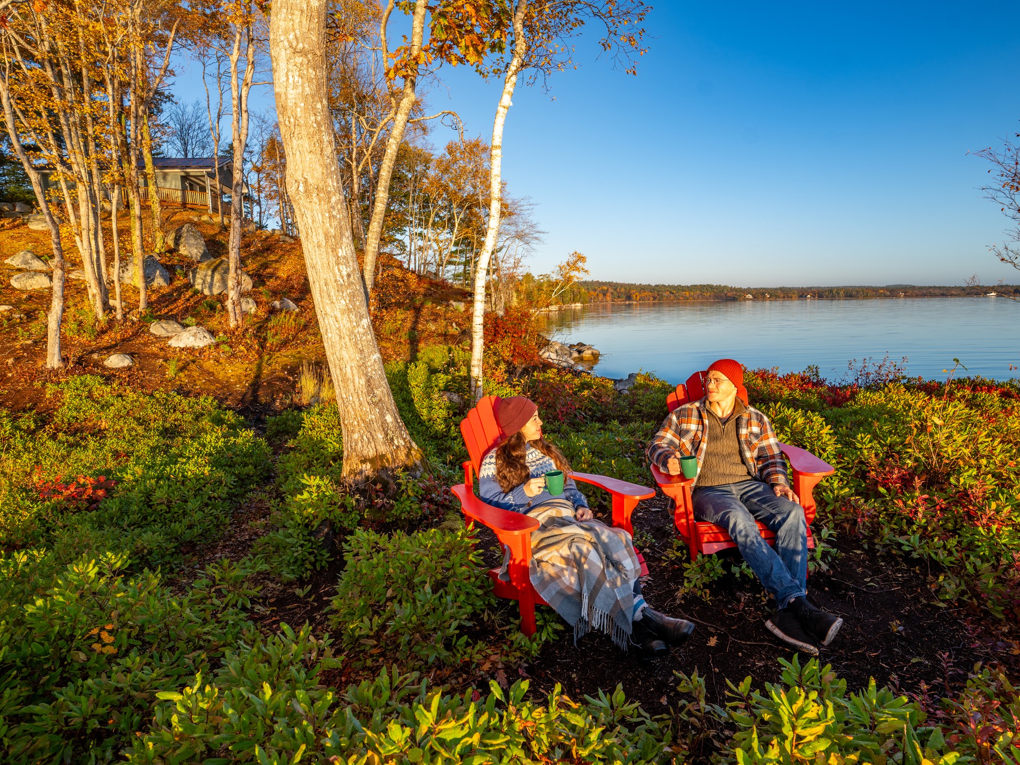 Two people enjoy coffee sitting in red Adirondack chairs next to the water at Boulder Cove Cottages in Shelburne, Nova Scotia.