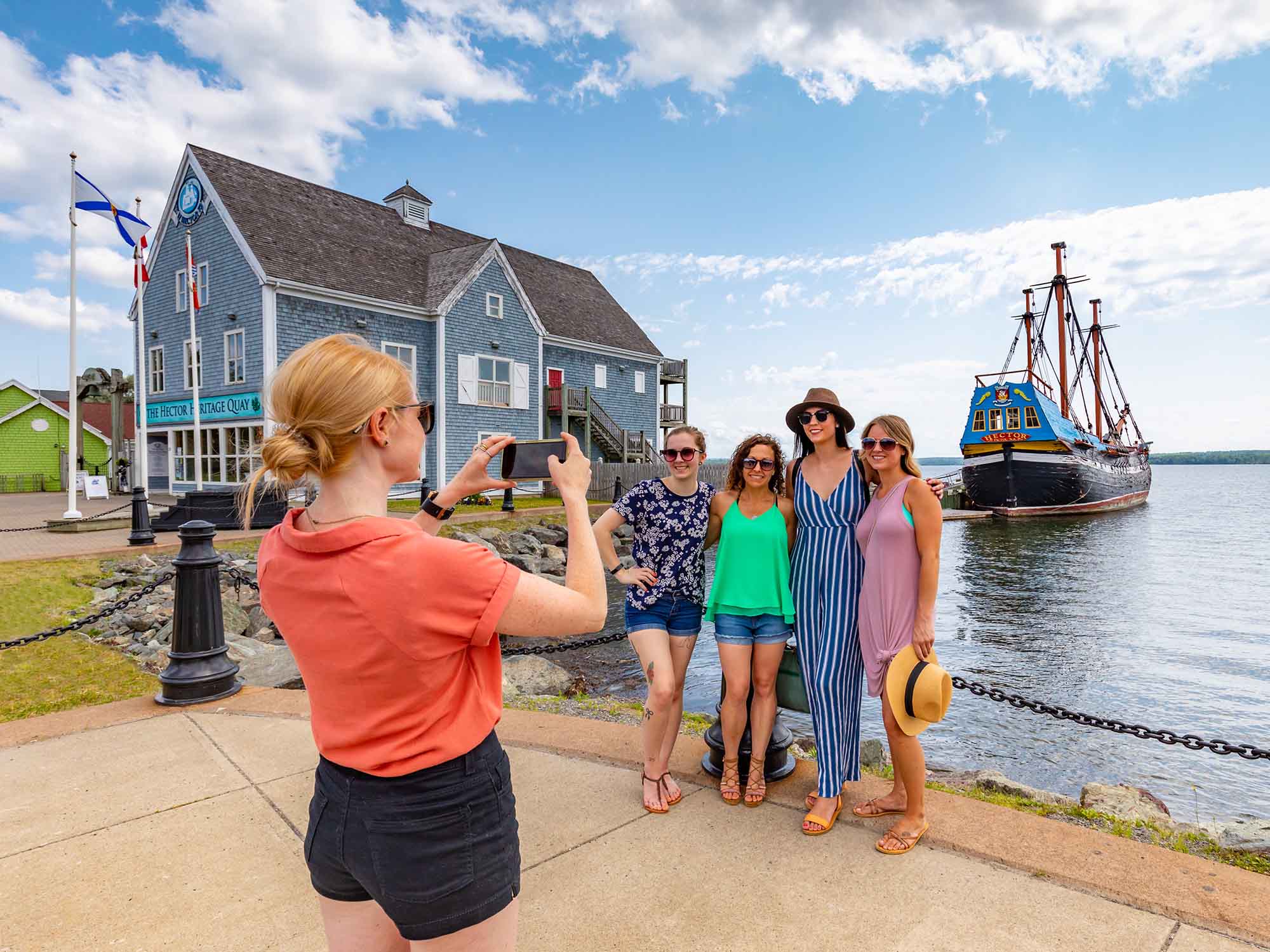 Friends getting their photo taken on the Pictou Waterfront with the replica of the Ship Hector and the Hector Heritage Quay museum behind them on a warm sunny summer day in Nova Scotia.