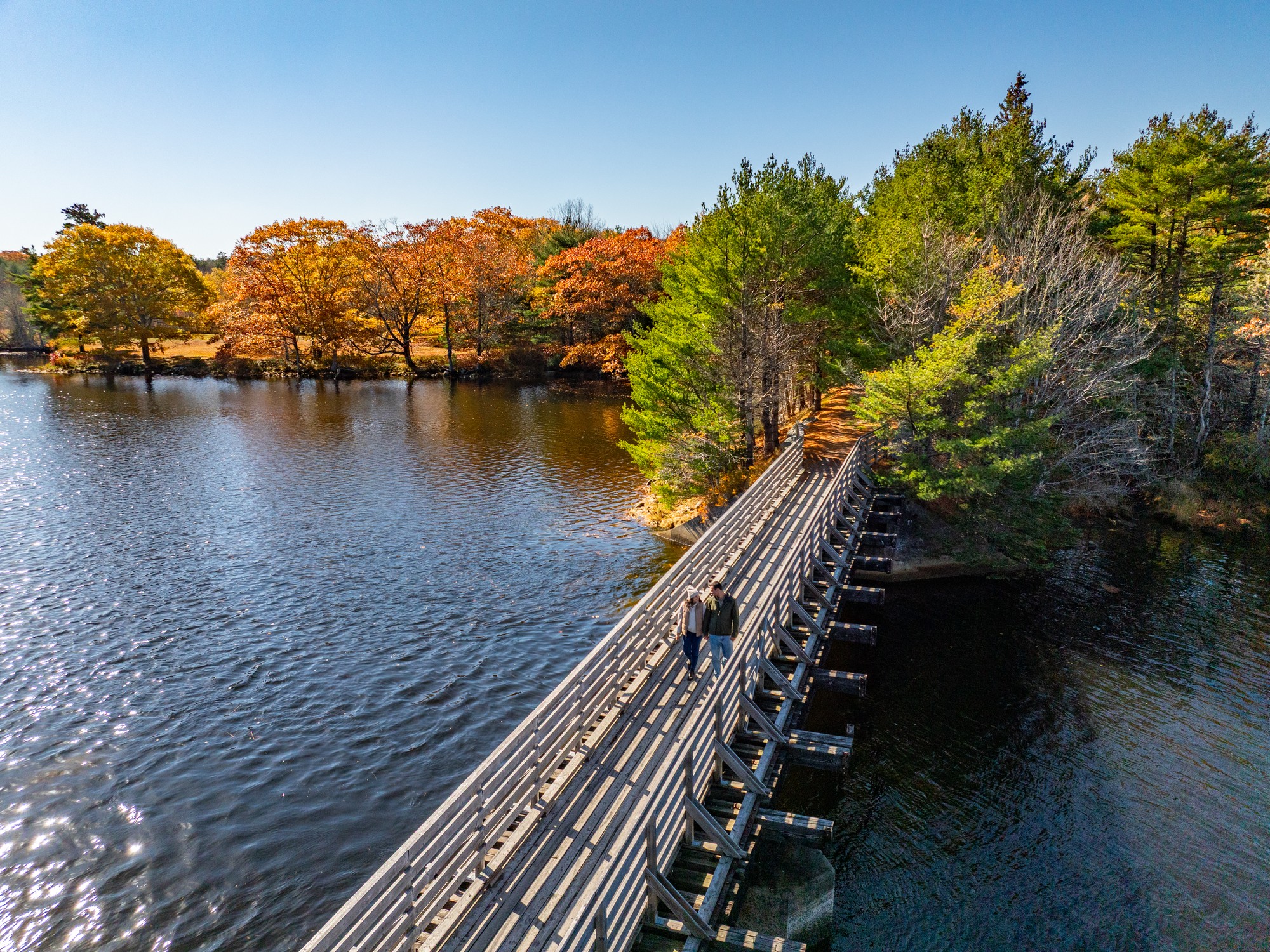 Two people walk across a bridge crossing a river on the Tom Tigney Trail in autumn near Shelburne, Nova Scotia