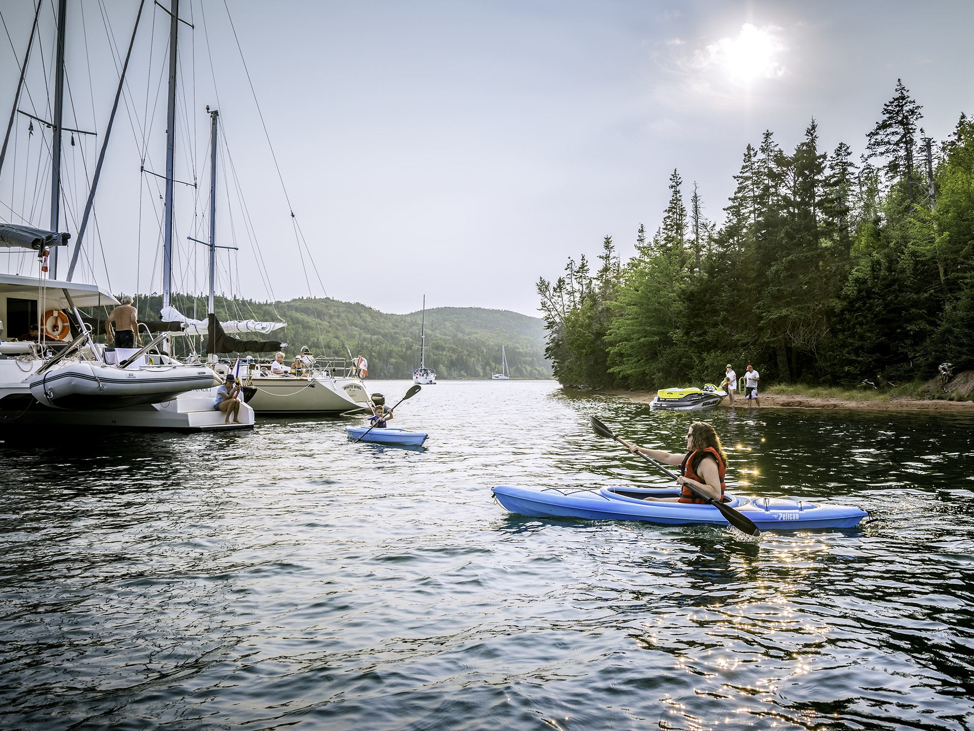 A person kayaking near two yachts that are anchored near Kidston Island in Baddeck on the Bras d'Or Lake in Cape Breton, Nova Scotia