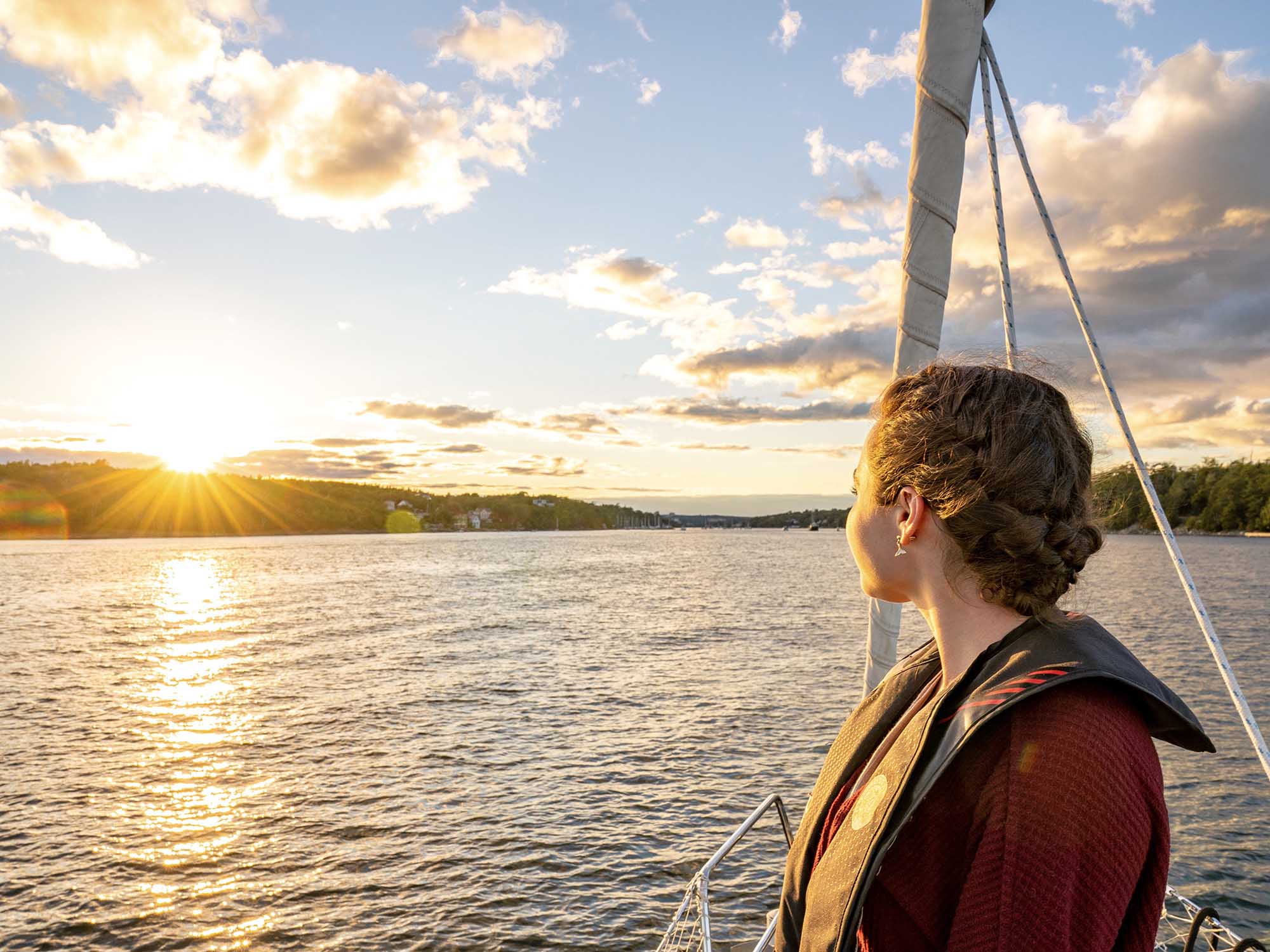 Person with a life vest on, standing near the front of a yacht sailing in Halifax's Northwest Arm, facing towards the summer sunset on a warm summer evening in Nova Scotia.