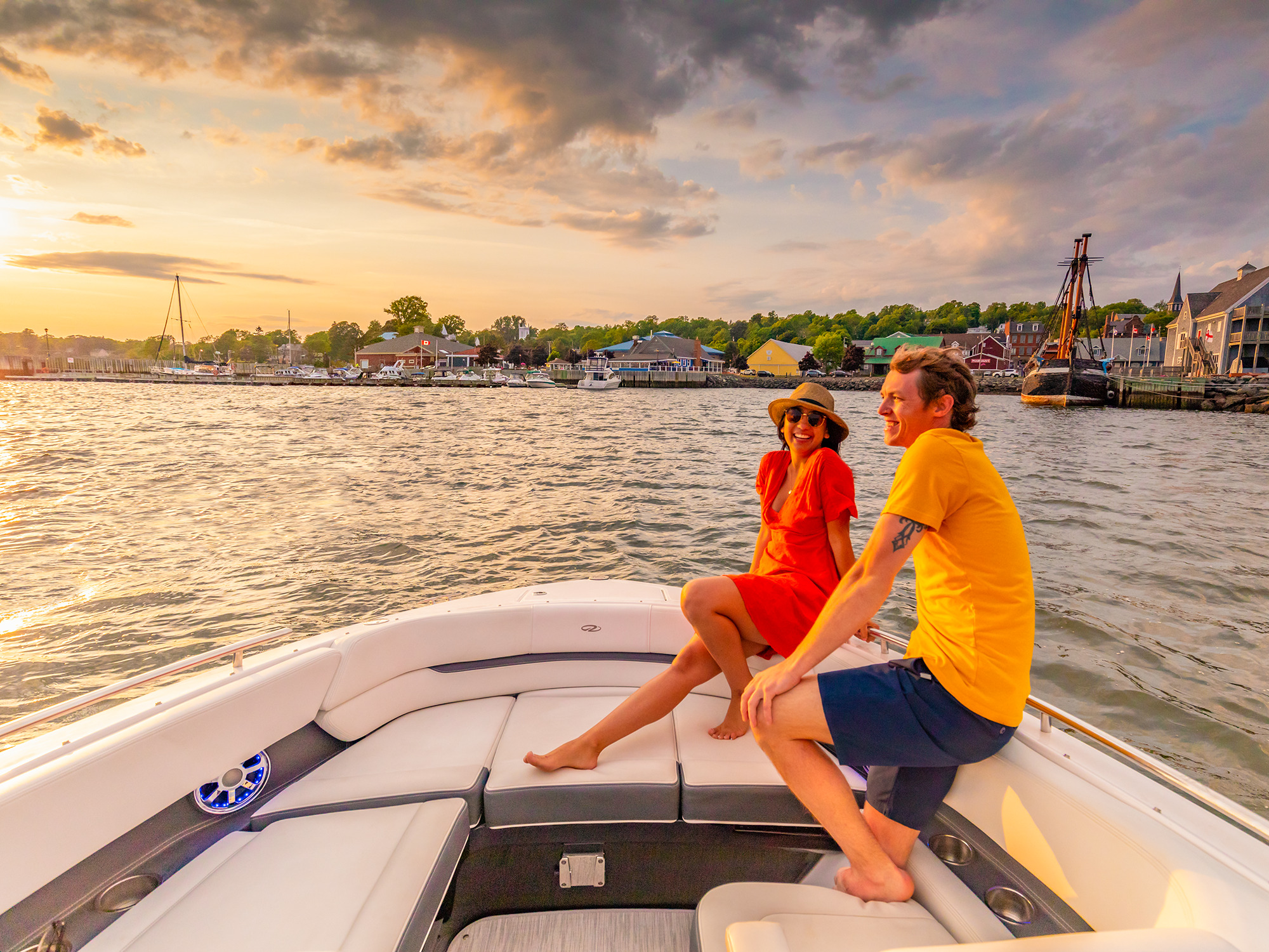 Two people smiling at each other as they sit together at the front of a boat in Pictou Harbour as the sun is setting over the Pictou Waterfront on a warm summer evening in Nova Scotia.