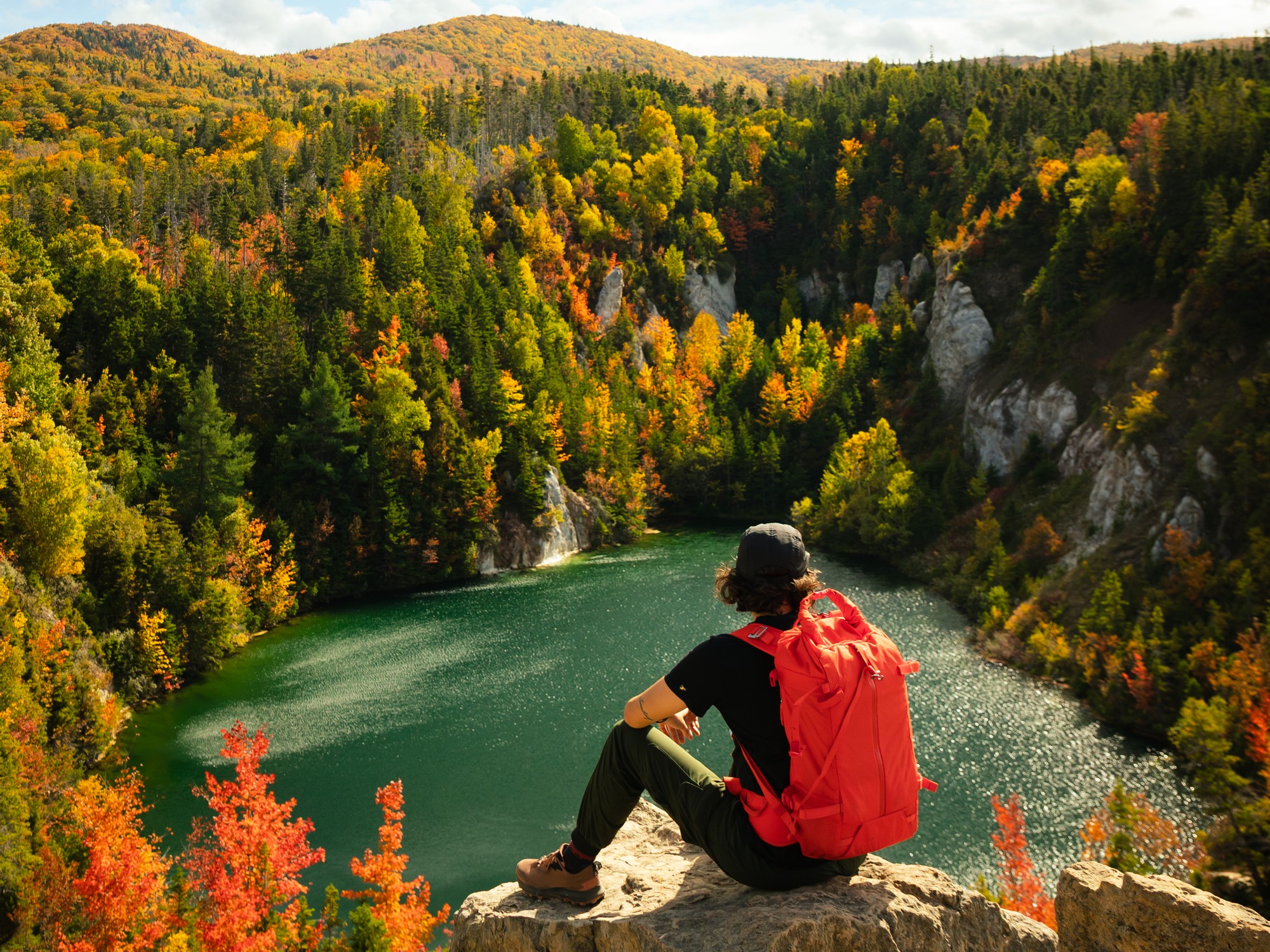 A person sits on a rock overlooking a lake at the bottom of a quarry with fall colours in the background in Cape Breton, Nova Scotia