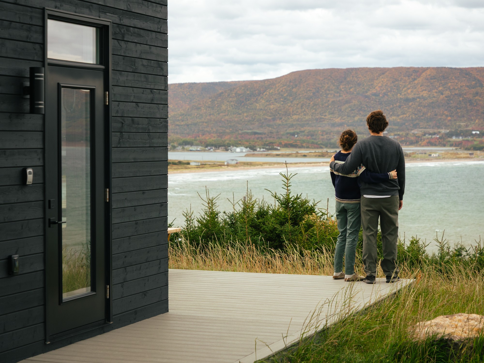 A couple admires the view from their waterfront cottage with fall colours in the background in Cheticamp, Nova Scotia