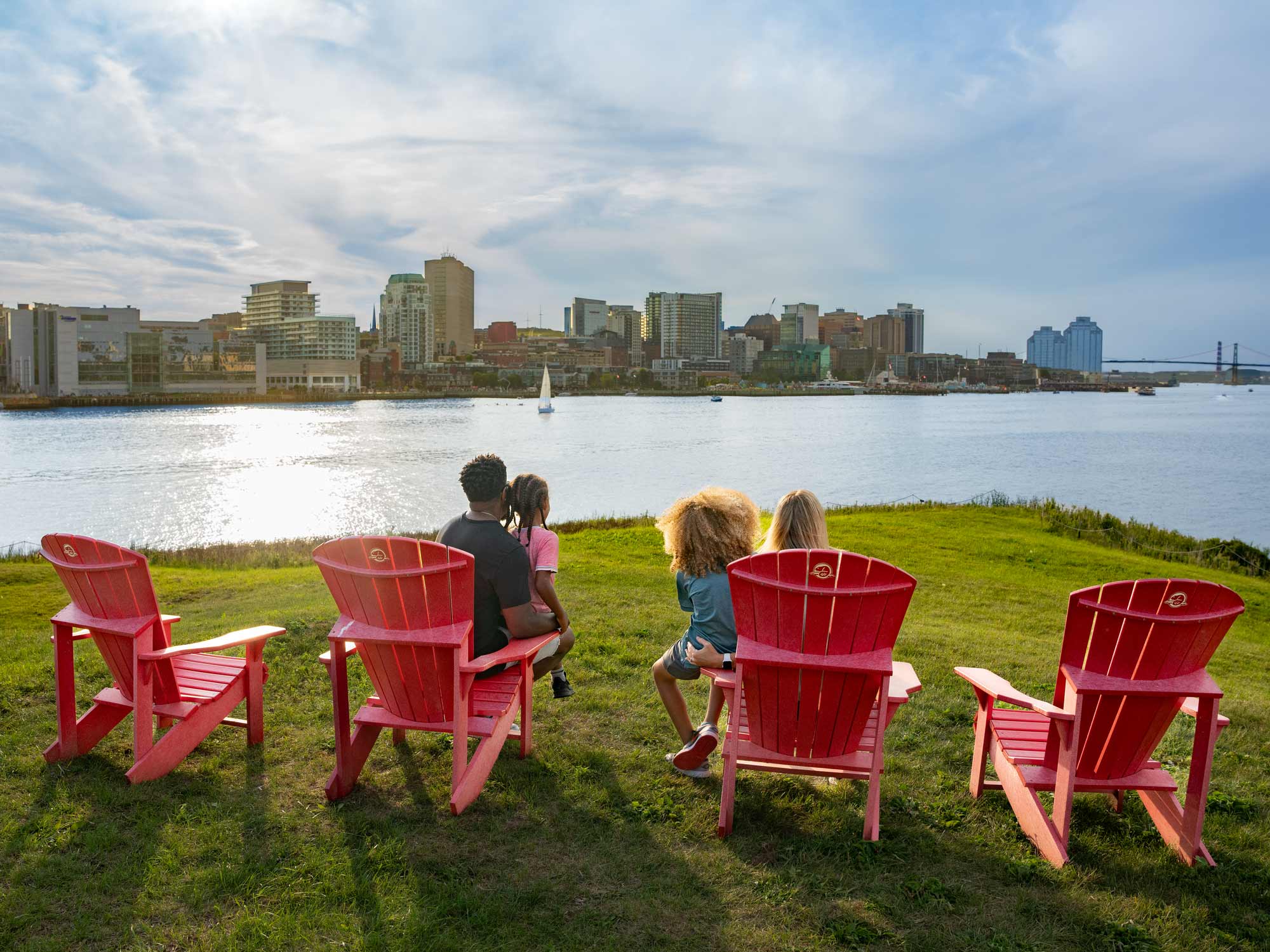 Parents and two children sitting on the Parks Canada red chairs on Georges Island National Historic Site in Halifax Harbour, looking out towards the Halifax Waterfront on a late afternoon in the summer in Nova Scotia