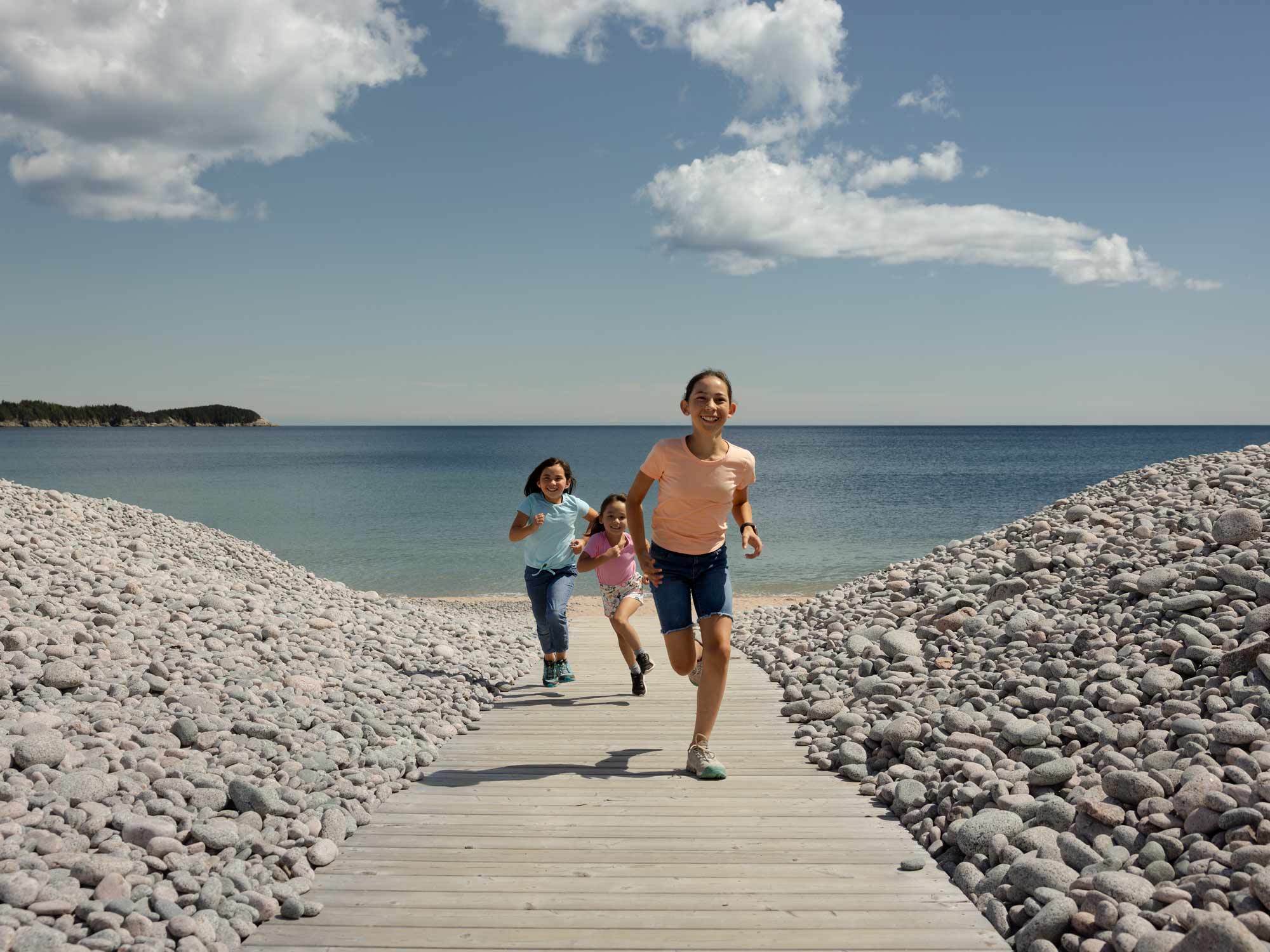 Three children running up the boardwalk from the sandy beach past the cobblestone that separated the beach from the parking lot at Ingonish Beach in Ingonish, Cape Breton, Nova Scotia.
