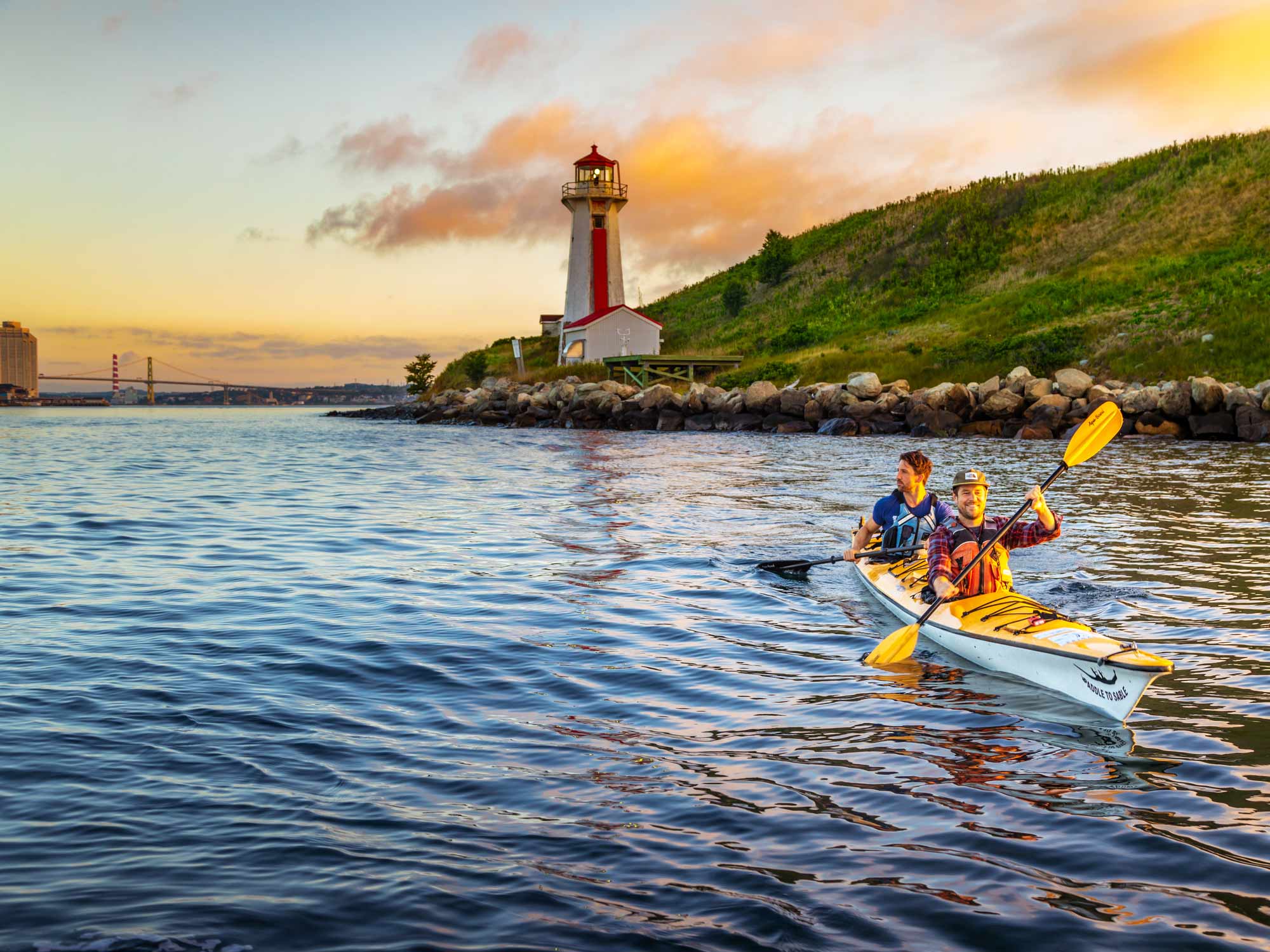 Two people kayaking near Georges Island National Historic Site in Halifax Harbour with the Halifax waterfront and MacDonald Bridge in the distance at sunset in Nova Scotia.
