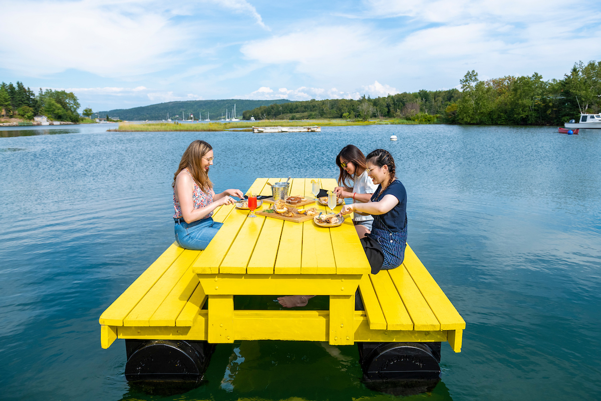 A group of people dine on a floating yellow picnic table at Inverary Resort in Baddeck, Nova Scotia