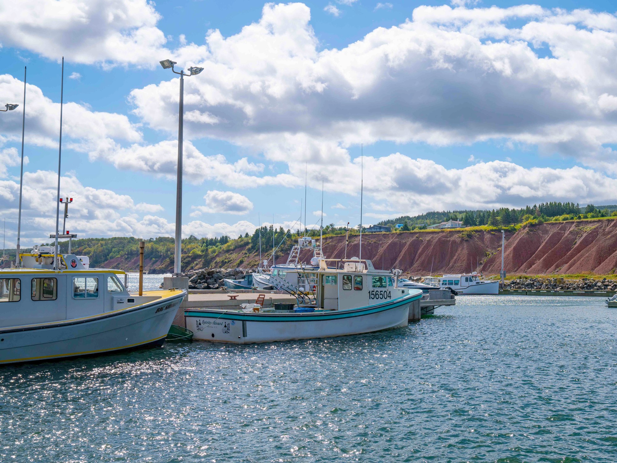 Fishing boats are docked at a wharf against a backdrop of red cliffs at Ballantyne's Cove, Nova Scotia