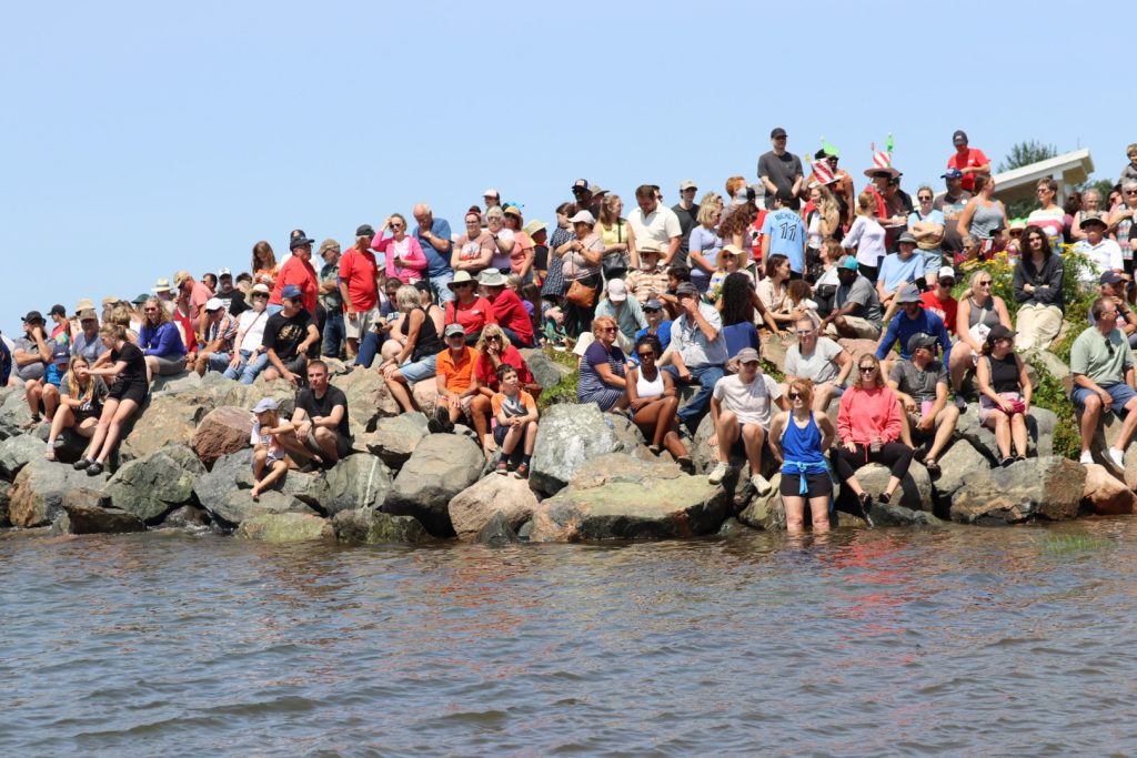 A very large group of people sits on rocks alongside the water watching an event.