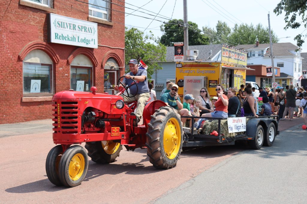 An older man sits on a red tracker pulling a group of people in a hay wagon during a parade.