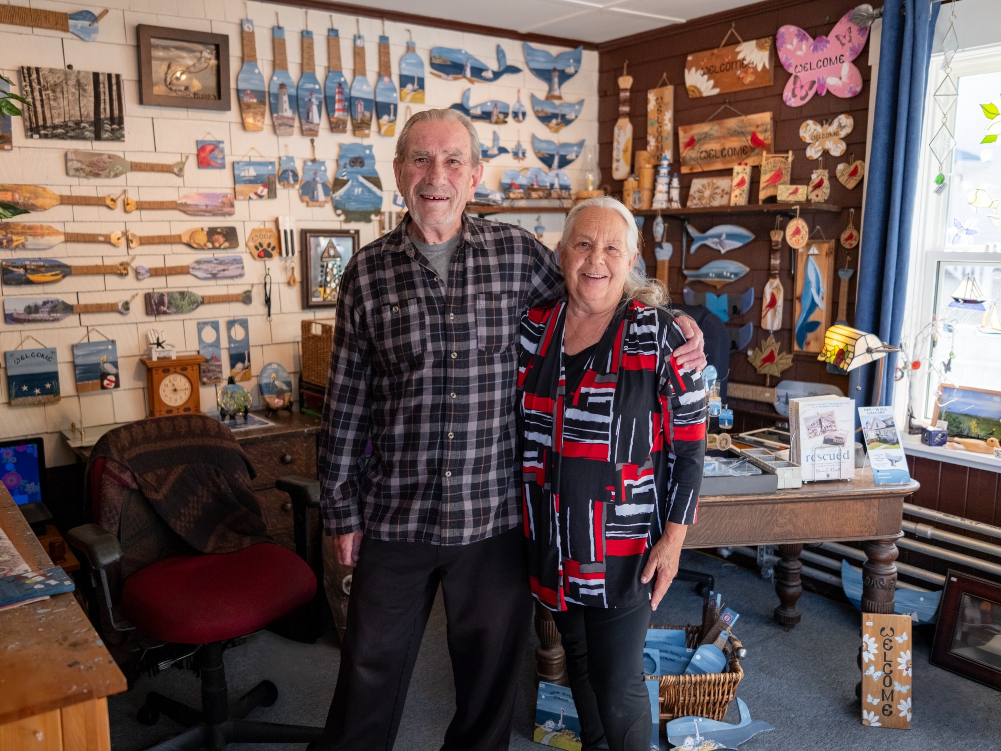 Two people have arms around eachother standing in front of a colourful wall of art at Off the Wall Gallery near Digby, Nova Scotia