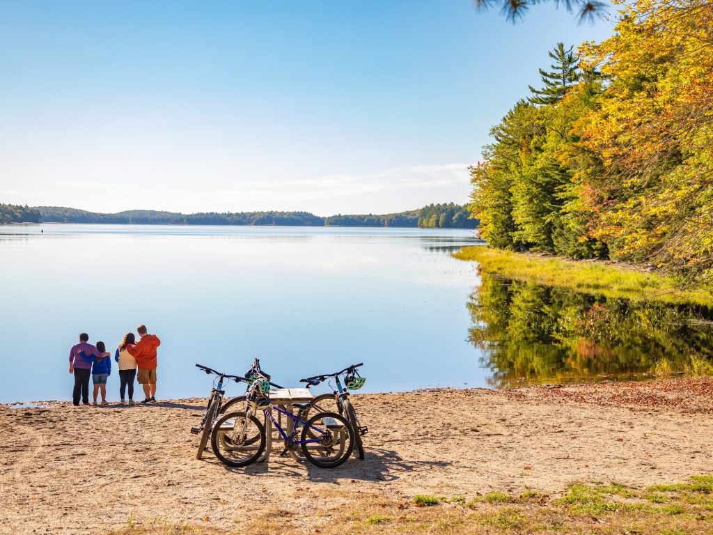 A family stands at a still lake with their bikes resting near by during autumn at Kejimkujik National Park and National Historic Site in Nova Scotia