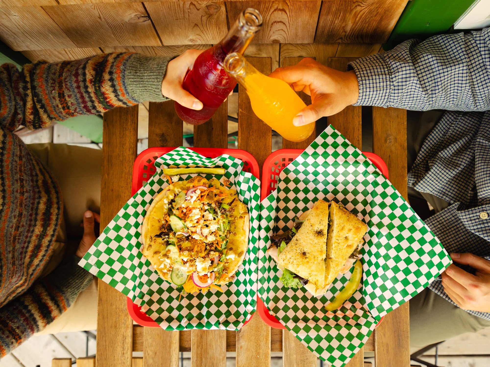 An overhead view of people's lunch of sandwiches. They are also doing a cheers with two soda bottles at Last Chance Sandwich in Cheticamp, Nova Scotia